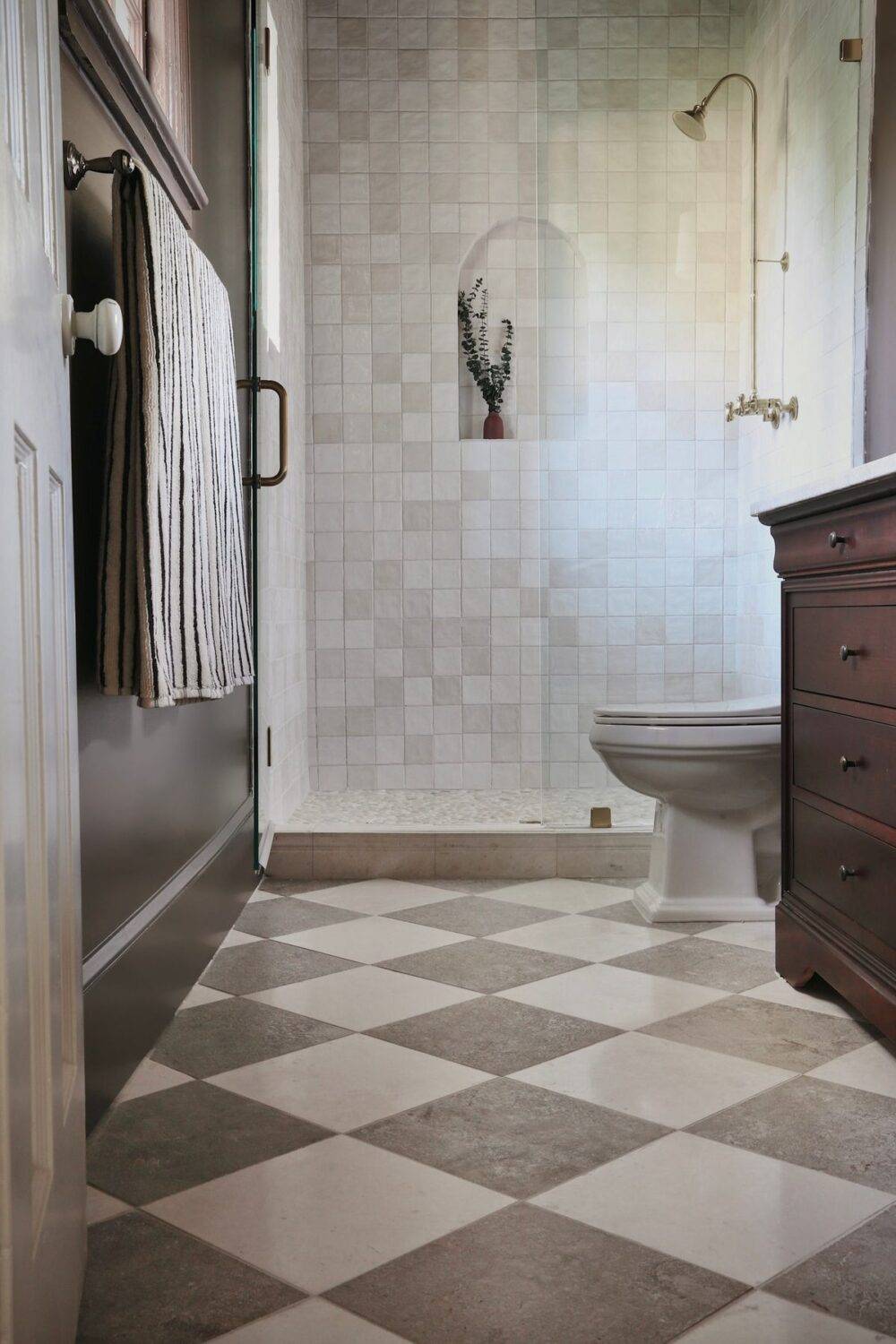 This elegant bathroom features handmade-look shower tile and a white and grey stone-look checkerboard tile floor. 